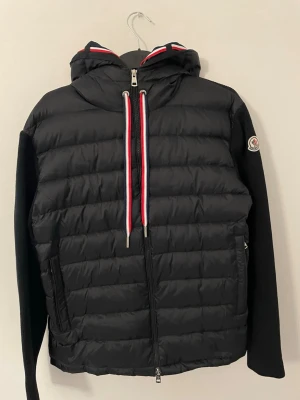 Moncler Cardigan  - Säljer en svart pufferjacka från Moncler med huva och coola rödvita detaljer på dragkedjan och luvan. Klassisk Moncler-logga på ärmen och två fickor framtill.