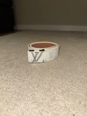 Vitt bälte med monogrammönster & silver spänne - Vit bälte med ikoniskt monogrammönster och svart metallspänne. Stilren och lätt att matcha till både vardag och fest. Snygg detalj som lyfter vilken outfit som helst.