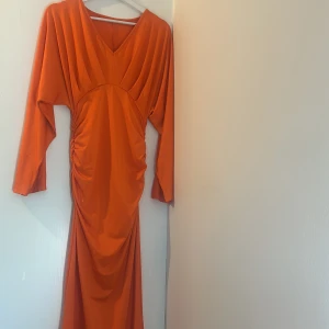 Lång orange klänning med v-ringning - Snygg långklänning i stark orange färg med v-ringad hals och långa ärmar. Klänningen har draperade detaljer över byst och midja samt rynkade sidor som ger en cool siluett. Perfekt för dig som vill sticka ut med en färgstark look.