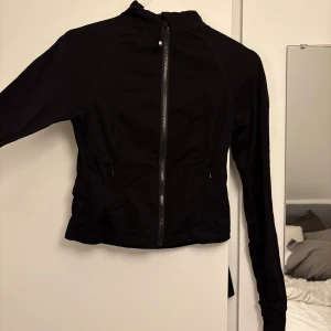 Svart träningskofta - Svart, figursydd vindjacka med hel dragkedja framtill och höga krage. Jackan har långa ärmar och två fickor med dragkedja. Perfekt för en aktiv stil och enkel att matcha med det mesta.