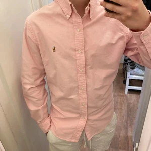 Polo Ralph Lauren (Rosa skjorta) - Säljer en klassisk rosa skjorta från Polo Ralph Lauren med button-down krage och broderad logga på bröstet. Skjortan är oanvänd och redo för en ny ägare! Perfekt för en clean och stilren look. Den är storlek S men passar även M!