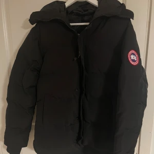 Svart dunjacka från Canada Goose - Säljer en svart dunjacka från Canada Goose med klassisk patch på ärmen. Jackan har huva, dragkedja och knappar framtill samt stora fickor. Perfekt för kalla dagar och riktigt snygg streetstil.