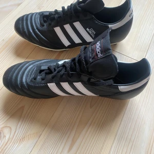 Adidas Copa Mundial fotbollsskor svart/vit 44 2/3 - Klassiska Adidas Copa Mundial fotbollsskor i svart skinn med vita detaljer och de ikoniska tre ränderna på sidorna. Skorna har snörning och fasta dobbar för gräsplan. Perfekta för dig som vill ha en tidlös och stilren look på planen.