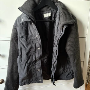 Mörkgrå jacka med stickade ärmar - Snygg svart jacka med stickade ärmar och krage. Jackan har dragkedja och knappar framtill samt en bröstficka med knapp. Kan va både drainer o Sthlm stil absolut🕺