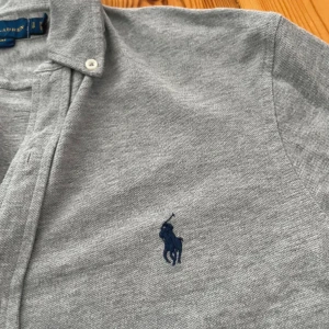 Grå Skjorta Ralph Lauren - Säljer en stilren grå långärmad skjorta från Ralph Lauren i mjuk bomull. Skjortan har klassisk button-down krage, vita knappar och den ikoniska mörkblå broderade loggan på bröstet. 
