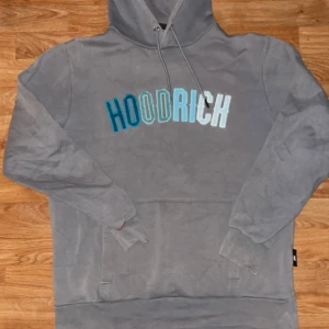 Grå hoodie från Hoodrich med brodyr - Grå hoodie från Hoodrich i storlek M med broderad logga i blå och turkosa nyanser över bröstet. Tröjan har huva med snörning och en klassisk känguruficka framtill. Mjuk insida i fleece och långa ärmar. Perfekt för chill dagar eller streetwear-stil.
