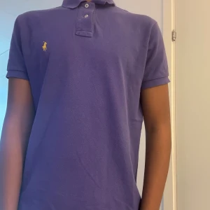 Lila pikétröja från Ralph Lauren S - Säljer en klassisk lila pikétröja från Polo Ralph Lauren i modellen Custom Fit. Tröjan har korta ärmar, krage med två knappar och den ikoniska gula loggan broderad på bröstet. Perfekt för en clean och stilren look.