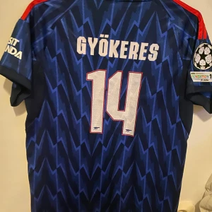 Arsenal blå bortatröja Gyökeres #14 - Säljer en Arsenal bortatröja från Adidas med Gyökeres och nummer 14 på ryggen. Tröjan är blå med blixtmönster, röda axelband och vita loggor. Champions League-märke på ärmen och 'Emirates Fly Better' tryck på bröstet. Perfekt för fotbollsfans!