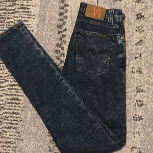 Mörkblå jeans från Nudie Jeans - Säljer ett par mörkblå jeans från Nudie Jeans med klassisk femficksdesign och snygg tvätt. Jeansen har raka ben och subtila slitningar för en cool look. Tillverkade i bomull och har Nudie Jeans logga på bakfickan och läderpatch i midjan.