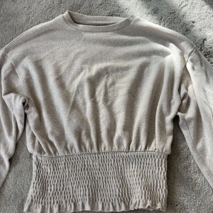 Beige långärmad tröja med smock - Superfin beige långärmad tröja med bred smockad midja och ärmslut. Tröjan har en rund halsringning och är gjord i ett mjukt material som är väldigt skönt mot huden. Tröjan sitter tajt runt midjan vilket ger en fin form på kroppen