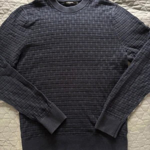 Mörkblå tröja från J.Lindeberg - Säljer en mörkblå tröja från J.Lindeberg med diskret ribbat mönster över hela plagget. Tröjan har rund halsringning, långa ärmar och breda muddar vid ärmslut och nederkant. Perfekt för dig som gillar stilrena och klassiska plagg.