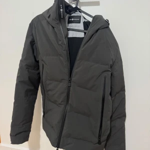 Svart dunjacka Sail Racing S - Säljer en grå dunjacka från Sail Racing i storlek S. Jackan har huva, två snedställda dragkedjefickor och är vadderad med 700 fill power dun. Snygg och stilren design med logga på kragen och insidan. Perfekt för kalla dagar. 