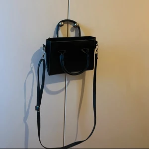 Svart axelväska i skinn med dragkedja - Snygg svart axelväska i skinn med både handtag och justerbar axelrem. Väskan har en stilren, fyrkantig form och ett stort huvudfack med dragkedja i mitten. Perfekt för dig som vill ha en enkel men trendig väska till vardags.