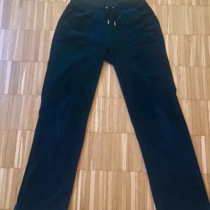 Mörkblå mjukisbyxor Juicy Couture - Mörkblå mjukisbyxor från Juicy Couture med raka ben och dragsko i midjan. Byxorna har fickor både fram och bak med snygga metalldetaljer och logga på snörändarna. Tillverkade i mjuk sammet för en riktigt skön känsla.
