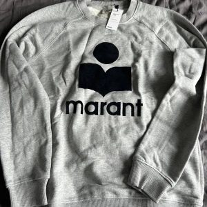 Grå sweatshirt från Isabel Marant - Grå sweatshirt från Isabel Marant med svart logotyptryck framtill. Klassisk rund halsringning, ribbade muddar och mjuk insida. Perfekt för en chill och stilren look. Tillverkad i bomullsmix för skön komfort. Den är tidigare artikelverifierad på Vinted. Skriv gärna om ni har frågor