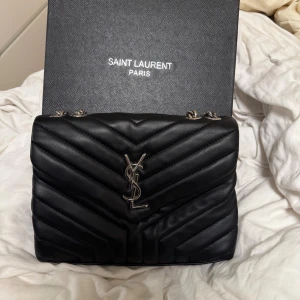 Svart quiltad handväska från Saint Laurent - Snygg svart handväska från Saint Laurent med quiltad design och silverfärgad YSL-logga framtill. Väskan har kedjerem med skinnparti, magnetlås och flera fack inuti, inklusive ett med dragkedja. Perfekt storlek för det viktigaste och riktigt lyxig vibe.