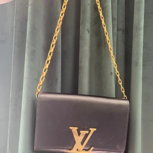 Svart Louis Vuitton axelväska i skinn - Snygg svart axelväska från Louis Vuitton med guldigt LV-spänne och kedja. Väskan är tillverkad i slätt skinn och har en stilren, rektangulär form. Perfekt för dig som gillar exklusiva accessoarer med lyxig känsla och ikonisk design.