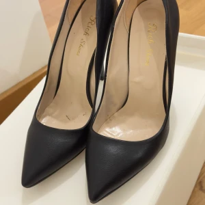 Svarta pumps med spetsig tå, stl 38 - Klassiska svarta pumps från Rich Shoes i storlek 38. Skorna har spetsig tå, hög smal klack och är tillverkade i ett slätt material som liknar skinn. Perfekta för dig som vill ha en stilren och elegant look.
