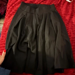 Svart kortkjol från Anna Field - Säljer en svart kortkjol från Anna Field med bred midja och snygga veck framtill. Kjolen har en klassisk A-linjeform och är perfekt för dig som gillar en stilren look. Materialet känns mjukt och följsamt, och kjolen är lätt att matcha med olika toppar.