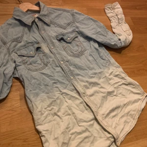 True Religion jeansskjorta blåvit - Snygg jeansskjorta från True Religion med cool dip dye-effekt i blått och vitt. Klassisk westernkrage, två bröstfickor med logodetaljer och tryckknappar framtill. Långärmad modell i bomull, perfekt för en avslappnad streetstil. Dock ett hål på ryggen, kan säkert sy ihop av nån händig.                                                Skick 5/10 