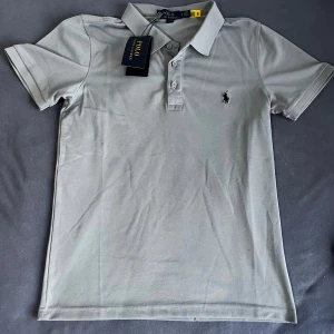 Ljusgrå pikétröja från Polo Ralph Lauren - Snygg ljusgrå pikétröja från Polo Ralph Lauren med klassisk krage och knäppning framtill. Tröjan har korta ärmar och den ikoniska broderade loggan på bröstet. Perfekt till jeans eller shorts för en clean och stilren look.