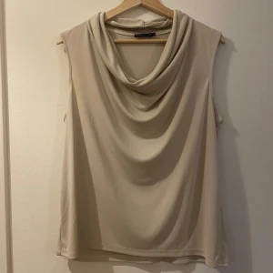 Offwhite ärmlös topp - Säljer en stilren offwhite/beige ärmlös topp från Stockh LM. Toppen har en snygg draperad krage och är tillverkad i ett mjukt, lätt material som faller fint. Passar under kavaj eller som elegant vardagstopp. 