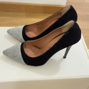 Svarta pumps med glittrig spetsig tå - Snygga pumps från High K.C. med svart mockaliknande material och glittrig, silverfärgad spetsig tå täckt av små stenar. Skorna har hög klack och elegant form, perfekta för att sticka ut. Insidan är beige och klacken är smal och svart. Se framsida av ena pumpens fattas de några stenar. 