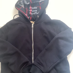 Burberry zip svart - Svart hoodie från Burberry med klassiskt rutigt foder i huvan och silverfärgad dragkedja. Tillverkad i 100% bomull, har huva med snörning och diskret broderad logga på bröstet. Perfekt för en chill och stilren look.