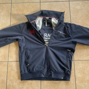 Marinblå vindjacka Sail Racing L - Snygg marinblå vindjacka från Sail Racing med broderade detaljer och amerikansk flagga på ryggen. Har en dragkedja framtill och två fickor med dragkedja. Perfekt till hösten men funkar även på vintern. Inga skador eller sådant på den. 