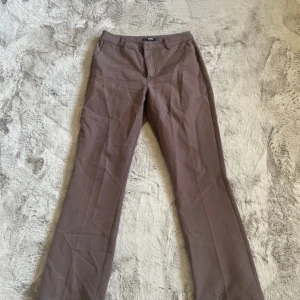 Bruna kostymbyxor med bootcut - Snygga bruna kostymbyxor med bootcut-snitt och klassisk passform. Byxorna har hällor för bälte och stängs med dragkedja och knapp. Perfekta för en stilren look och passar till många olika outfits.