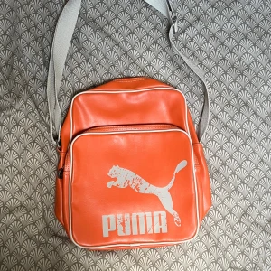 Orange axelväska från Puma - Cool orange axelväska från Puma med vit logga och detaljer. Väskan har justerbar grå axelrem, stort huvudfack och en praktisk ytterficka med dragkedja. 👍🏼