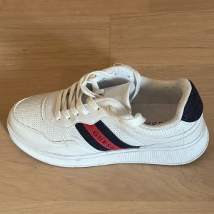 Vita sneakers från Guess strl 43 - Snygga vita sneakers från Guess med klassiska röda och marinblå ränder på sidan. Skorna har snörning, perforerade detaljer och en platt, vit sula. Tillverkade i syntet och textil för en fräsch och sportig look. Perfekta för dig som gillar stilrena och trendiga sneakers.