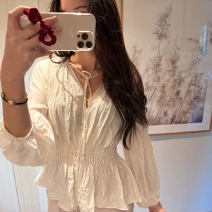 Vit blus med knyt och volang - Säljer denna super fina vita blus med gulddetaljer på snörena. Blusen är figursydd i Miriam och vid i ärmarna. Den är helt oanvänd och har prislappen kvar, heller inga defekter. 