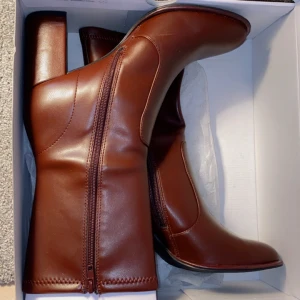 Bruna boots med klack och dragkedja - Snygga bruna boots i skinnimitation med fyrkantig klack och dragkedja på sidan. Skorna har en rund tå och stilren design som passar perfekt till höst- och vinteroutfits. Perfekta för dig som vill ha en klassisk men trendig look.