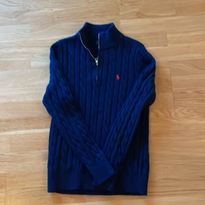 Marinblå stickad tröja Polo Ralph Lauren - Säljer en marinblå kabelstickad tröja från Polo Ralph Lauren med halv dragkedja och röd broderad logga på bröstet. Tröjan har hög krage och är långärmad, tillverkad i ullblandning för extra värme och stil. Storlek M men passar också S och är 170-180 