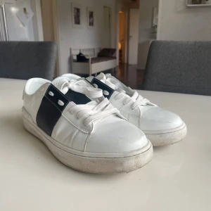 Valentino Garavani vita sneakers - Snygga vita sneakers från Valentino Garavani med bred svart läderdetalj över sidan och coola nitar baktill. Klassisk rund tå, vita snören och platt sula. Perfekta för dig som vill ha en stilren men edgy look.