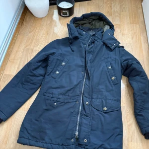 Mörkblå parkas från Svea, storlek S - Säljer en mörkblå parkas från Svea med snygga detaljer som stora fickor med knappar, dragkedja och huva. Jackan är fodrad med olivgrönt innerfoder och har ribbade muddar vid ärmsluten. Perfekt för kalla dagar och riktigt skön att bära.