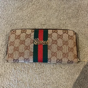 plånbok med mönster och ränder - Snygg plånbok med klassiskt mönster i beige och brunt. Framsidan har gröna och röda ränder samt ett guldfärgat . Plånboken har dragkedja med logga och är tillverkad i canvas med detaljer i skinn. Endast swish betalning skickas spårbart 59kr