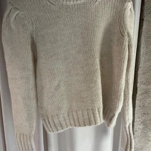 Beige stickad tröja från ADPT - Mysig beige stickad tröja från ADPT med rund halsringning och ribbade muddar. Tröjan har långärmade ärmar med lätt puff vid axlarna för en trendig look. Perfekt för lager-på-lager under kyliga dagar.