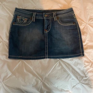 True Religion jeanskjol med kontrastsömmar - Säljer en snygg blå jeanskjol från True Religion med tydliga vita kontrastsömmar och klassiska bakfickor med broderad logga. Kjolen har bälteshällor, knapp och dragkedja framtill. Perfekt för dig som gillar streetstyle och denim.