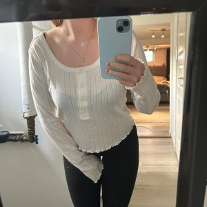 Vit ribbad långärmad tröja med knappar - Säljer en söt vit långärmad tröja med ribbat mönster och knappar framtill. Tröjan har en rundad halsringning och små volangdetaljer vid ärmslut och nederkant. Perfekt för lager-på-lager och chill vardagsstil.🩷