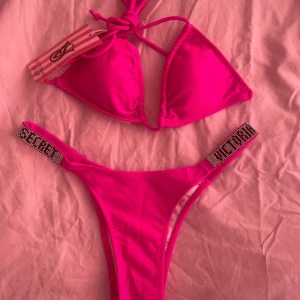 Rosa bikini från Victoria's Secret - Snygg rosa bikini från Victoria's Secret med trekants-bh och knyt i nacken. Underdelen har glittriga detaljer med texten 'SECRET' på sidorna. Perfekt för stranden eller poolen, riktigt poppig färg och trendig design.