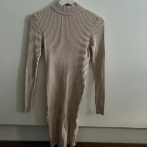 Beige ribbad långärmad klänning - Ribbad beige klänning med lång ärm och hög hals. Modellen är kroppsnära och har en stilren, enkel design som passar perfekt för dig som gillar minimalistiskt mode. Materialet är stretchigt och mjukt, vilket gör den både bekväm och snygg.