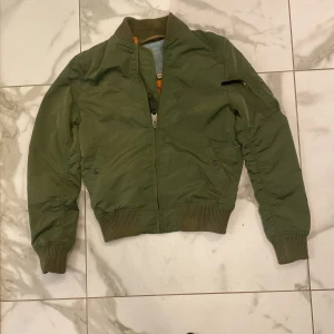 Grön bomberjacka från Alpha Industries - Säljer en klassisk grön bomberjacka från Alpha Industries med orange foder. Jackan har ribbade muddar, dragkedja framtill och fickor med tryckknapp samt en ärmficka med dragkedja. Perfekt för dig som vill ha en stilren och tidlös look.