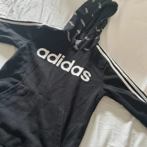 Svart Adidas hoodie med logga i storlek 152 - Svart hoodie från Adidas i storlek 152 med stor vit logga framtill och klassiska vita ränder längs ärmarna. Huva med Adidas-tryck och stor magficka. Perfekt för en sportig och avslappnad stil.