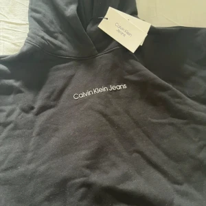 Svart hoodie från Calvin Klein Jeans i storlek 12 - Svart hoodie för barn/ ungdom Calvin Klein Jeans med vit broderad logga på bröstet. Klassisk huva och mjukt material, perfekt för en chill och stilren look. Passar dig som gillar enkel och snygg streetwear.