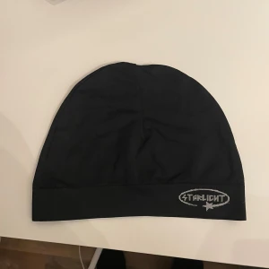 Svart mössa från Starlight - Svart, slimmad mössa från Starlight med grått broderat logotyptryck framtill. Enkel och stilren design. Tillverkad i mjukt och stretchigt material som sitter skönt på huvudet.