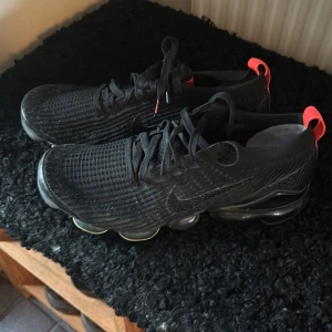 Nike Vapormax svarta sneakers - Svarta Nike Vapormax sneakers med stickad ovandel och coola genomskinliga sulor. Skorna har snörning och röda detaljer på hälen som ger en snygg kontrast. Perfekta för dig som gillar sportig och modern stil. Storlek 42,5, knappt använda. Ej originalsula i, annan Nike. 