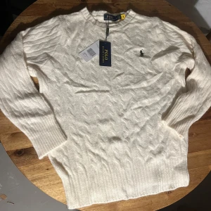 Vit kabelstickad tröja Polo Ralph Lauren - Kabelstickad vit tröja från Polo Ralph Lauren med klassisk rund hals och broderad logga på bröstet. Tröjan har långa ärmar och ribbade muddar samt nederkant. Perfekt för dig som gillar stilren och tidlös design. Storlek M men passar nog mer S. Aldrig använd, säljer för att den var för liten för mig