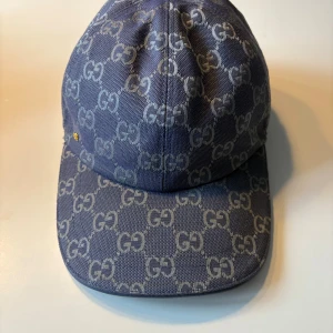 Blå Gucci keps med GG-mönster - Snygg blå keps från Gucci med ikoniskt GG-mönster över hela kepsen. Kepsen har böjd skärm och justerbar rem baktill i metall. Materialet är bomull och polyester, vilket gör den både bekväm och hållbar. Perfekt för dig som vill ha en lyxig streetstyle-look. Använd väldigt få gånger. Pris kan diskuteras vid snabb affär 
